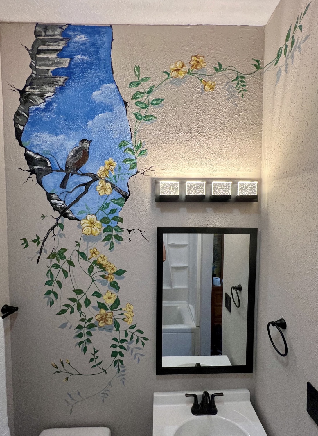 A bathroom mural with a bird perched among yellow blossoms beside an opening to a blue sky.